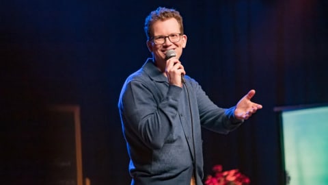 Popular internet educator and author Hank Green stands mid-gesture under vibrant stage lighting during a performance of his debut stand-up comedy special, Pissing Out Cancer, which premiered via Dropout Presents. Wearing his signature thick-rimmed glasses, a structured slate-blue buttoned cardigan polo, and casual tan trousers, he smiles broadly while holding a corded microphone close to his mouth with his right hand and extending his left hand outward to engage with the audience.