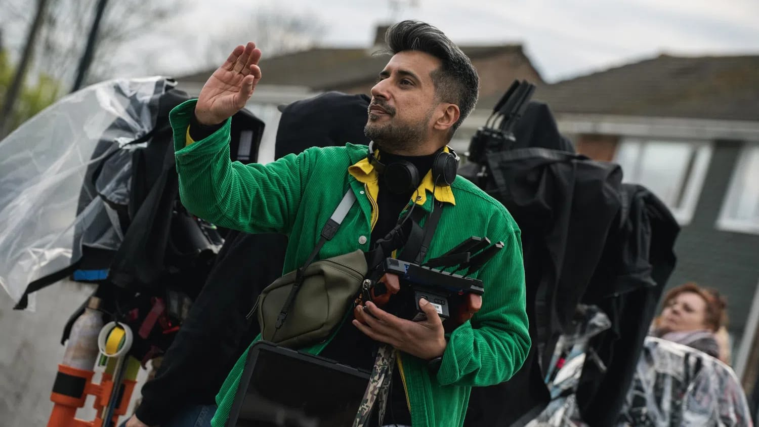 A medium shot of director Asad Abbasi gesturing with an open hand on the outdoor set of A Good Girl's Guide to Murder Series 2. He wears a vibrant green corduroy jacket over a yellow collared shirt and black turtleneck, equipped with production headphones around his neck, a shoulder bag, and a tablet. Cameras covered in black protective plastic wrap form a busy background alongside a crew member wearing a clear rain poncho.