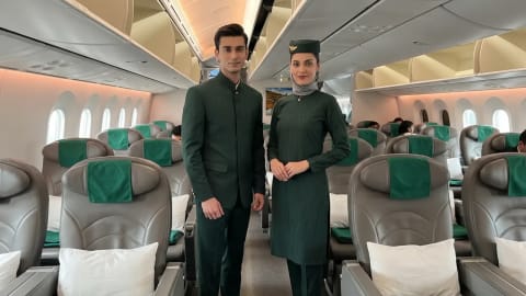 Two flight attendants posing inside an airplane cabin wearing deep green uniforms designed by HSY for PIA. An flight attendant team stands inside the cabin of a commercial aircraft, wearing deep green uniforms designed by HSY for Pakistan International Airlines (PIA). The man on the left wears a tailored mandarin-collar green jacket with matching trousers and polished black shoes, while the woman stands next to him wearing a structured green tunic with a matching cap, light-grey headscarf, straight pants, and black heels. They pose gracefully in the aisle between rows of grey business class seats fitted with green headrests, white pillows, and neatly folded green blankets, beneath a warm overhead lighting system.