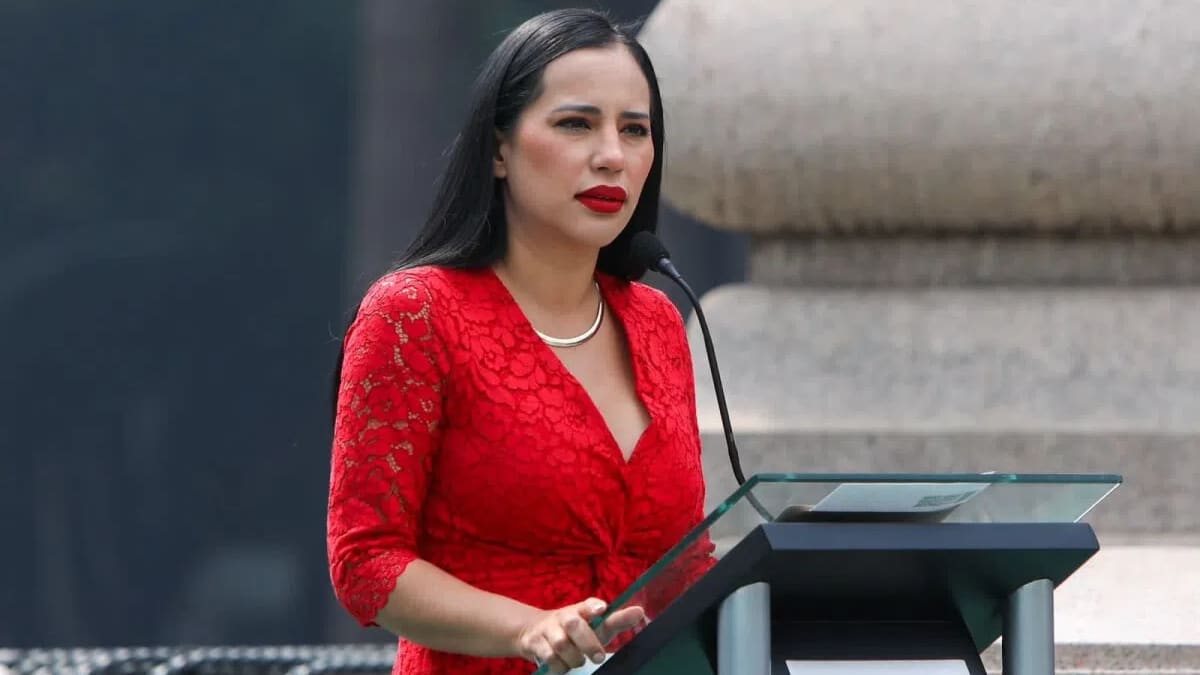 This image features Mexican politician Sandra Cuevas delivering a speech at a podium. She is wearing a striking scarlet red floral lace dress that has three-quarter length sleeves, a deep V-neckline, and a wrap-style bodice. Her look is complemented by bold red lipstick, straight dark hair, and a simple gold necklace. Cuevas stands behind a modern, clear-topped podium with two microphones, positioned in front of a large, light-colored stone structure. 