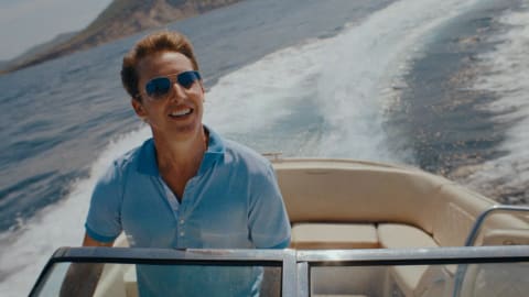 English singer-songwriter James Blunt smiles brightly while piloting a motorboat through deep blue open waters, leaving a massive white sea spray wake trailing behind him. He wears a light blue polo shirt with darker blue trim on the collar and sleeves, paired with classic metallic aviator sunglasses that reflect the coastal surroundings. Behind him, a rugged, mountainous coastline rises out of the ocean under a clear, bright sky, framing the sun-drenched, high-speed maritime scene.