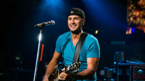 The image captures American country music singer and songwriter Luke Bryan performing live on stage during a concert. He is smiling warmly while looking slightly off-camera, dressed in a casual teal v-neck t-shirt and a backward black baseball cap featuring a white deer antler logo. Bryan is holding and playing an acoustic guitar, which is supported by a thick black strap slung over his shoulder. He stands positioned behind a silver microphone on a stand, illuminated by dramatic blue stage lighting that cuts through the dark background. The background features elements of a live concert setup, including hazy smoke effects and glimpses of stage equipment, creating an energetic and intimate performance atmosphere.