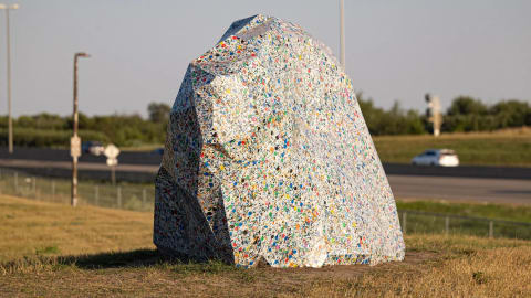 The image features "Modern Erratics," a controversial $297,490 permanent public art installation commissioned by the City of Saskatoon consisting of three large boulder-shaped sculptures crafted out of thousands of kilograms of compressed recycled plastic waste. Designed by the Calgary-based artist duo Sans façon to mimic glacial erratic boulders while making a statement on environmental pollution, this specific piece located in Diefenbaker Park is constructed from multi-colored recycled plastic bottle caps. The installation has sparked a heated public debate across Saskatchewan over tax spending, with some residents and the Canadian Taxpayers Federation mocking it as a waste of funds while city officials and arts advocates defend the project for promoting environmental awareness and community conversation.