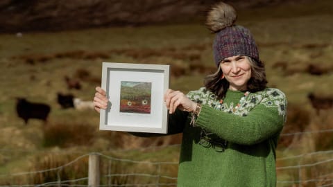A medium-shot photograph captures a fair-skinned woman standing outdoors in a rural, hilly landscape, smiling gently as she holds up a small, square, light-gray framed artwork. She is positioned on the right side of the frame, wearing a thick, vibrant green knit sweater with a white and dark green Fair Isle patterned yoke, a multi-colored knit beanie with a large faux-fur pom-pom, and dark-rimmed eyeglasses tucked into the front of her collar. Her shoulder-length brown and gray hair frames her face, and she wears long, colorful beaded necklaces. With both hands, she displays the framed piece of art, which depicts a colorful, abstract textile landscape featuring small sheep figures against a hillside, complete with a signature at the bottom. The background consists of a sloping, grassy brown and green hill under bright, natural daylight, with several dark sheep grazing in the distance behind a wire mesh fence with wooden posts that runs along the bottom foreground.