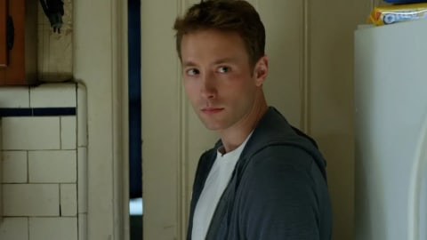 A medium close-up shot capturing actor Kent Moran as the character Jaden Miller from a side-profile angle as he looks over his left shoulder directly toward the camera. He has light brown, short-cropped hair styled slightly upward at the front, a fair complexion with a subtle reddish mark near his left eye, and a serious, guarded facial expression. He wears a plain white crewneck t-shirt underneath an unzipped, textured charcoal-grey hoodie jacket. The background depicts a dimly lit, weathered home interior, likely a kitchen entryway. To the left, a vertical section of a wall is visible, covered in square, off-white ceramic tiles divided by a prominent dark grout line. A sliver of an adjacent doorway reveals a dark blue background behind him. To the right, the flat white side panel of a refrigerator fills the frame, upon which a yellow package of Oreo cookies rests on top.