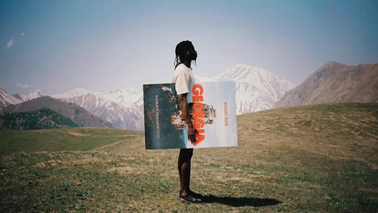 The image presents a wide, cinematic shot of a person standing sideways in a grassy field set against a backdrop of rolling green hills and large, snow-dusted mountains under a clear blue sky. The individual has long dark hair styled in braids or dreadlocks and wears a plain white shirt, dark shorts, and sandals. They are holding a large, rectangular poster or vinyl sleeve horizontally across their torso; the right side of the poster features the word "GEORGIA" written in large, bold red capital letters stacked vertically over a portrait of a face.