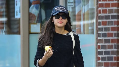 An attractive woman walks down a sidewalk in a city context, captured from a straight-on perspective. The individual, identified as singer-songwriter Olivia Rodrigo, steps forward on her left foot, looking off toward her right with a gentle, thoughtful expression. She wears a navy blue baseball cap with white embroidered text reading "SUPERVINYL," dark black sunglasses with thick frames, and her long brown hair flows freely down past her shoulders. Her casual ensemble consists of a loose-fitting black sweater layered over a black-and-white striped shirt, a pleated olive green knee-length skirt, and tall black leather boots that reach her mid-calf. In her right hand, she holds a yellow apple with a bite taken out of it, and a cream-colored canvas tote bag hangs from her left shoulder, with her left hand lightly holding onto a cell phone. The background consists of a brick building with a large glass window that features neon lights reflecting green and purple hues inside. A small printed sign is visible in the upper right corner of the window. The sidewalk is made of gray concrete blocks, transitioning to an asphalt street with white painted crosswalk lines in the immediate foreground. Olivia Rodrigo channels a chic, effortless street-style aesthetic while walking through New York City, wearing a relaxed black off-the-shoulder wrap sweater layered over a classic black-and-white striped top, paired with an olive green A-line midi skirt, knee-high black leather boots, a dark baseball cap, and oversized black sunglasses while casually holding a half-eaten apple.
