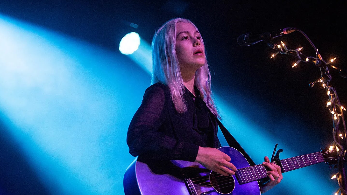 An eye-level shot captures singer-songwriter Phoebe Bridgers performing live on stage under intense blue and white spotlights. She has long, platinum blonde hair and wears a sheer black long-sleeved blouse with a high tie neck. She looks upward with an emotional expression while strumming a dark acoustic guitar with a prominent pickguard and a capo clamped on the neck. To her right, a black microphone stand is heavily wrapped in glowing, warm-toned fairy lights, creating a stark, moody contrast against the dark, hazy background of the venue.  Who is Phoebe Bridgers? Phoebe Bridgers (born August 17, 1994) is an American singer, songwriter, and musician known for her melancholic indie-folk and rock sound, confessional lyrics, and signature skeleton-suit stage attire. She has earned widespread critical acclaim and multiple Grammy nominations as a solo artist and as part of the indie supergroup Boygenius.