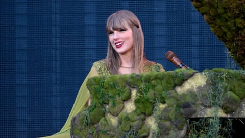 Taylor Swift sits smiling at a moss-covered grand piano on an outdoor stage during a concert performance. She is wearing a flowing, olive green gown with a matching cape and delicate lace detailing on the shoulders. Her blonde hair is styled straight with bangs, and she wears her signature bright red lipstick. The piano is heavily decorated with realistic green moss and hanging faux foliage, blending into a large mossy structure on the right. A wooden microphone stands near the piano keys. The background consists of a large, dark textured LED video screen showing a faint pattern.