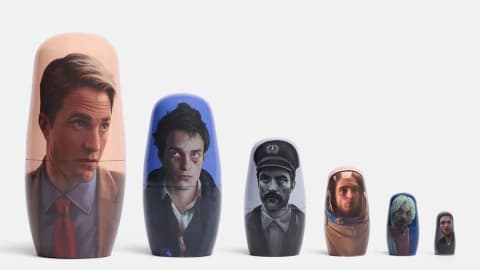 A set of six wooden nesting dolls decreases in size from left to right, each painted with a detailed portrait of actor Robert Pattinson representing a different film role. The largest doll features a clean-shaven Pattinson in a tan suit and red tie against a peach background. The second shows him looking disheveled with dark, messy hair and an untidy collared shirt against a blue background. The third doll is in black and white, depicting him with a thick mustache wearing a peaked captain's hat and a buttoned uniform jacket. The fourth doll portrays him looking upwards while wearing an astronaut helmet and suit. The fifth doll features him with messy, bleach-blond hair and a faint beard against a blue backdrop, and the smallest doll displays a close-up portrait of his face against a dark background.