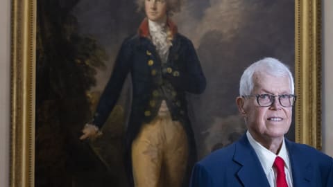 The image features a split focus between historical portraiture and a contemporary portrait: in the background, a large, gilt-framed oil painting depicts a young, aristocratic 18th-century gentleman with powdered hair, a high cravat, a dark tailcoat with bright buttons, and tan breeches, gesturing toward a dramatic, overcast landscape; standing prominently in the right foreground is an elderly Caucasian man with short, neat white hair, thin-framed glasses, and a subtle smile, dressed formally in a sharp blue suit jacket, a white collared dress shirt, and a solid red tie.