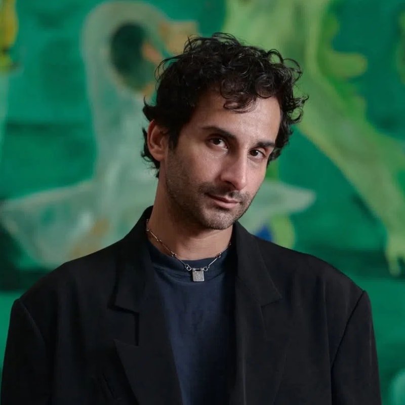 A close-up portrait shows Pakistani-American figurative artist Salman Toor, captured from the chest up while posing in front of one of his large, expressive oil paintings characterized by fluid forms and a dominant green and celadon color palette. Toor is dressed in a structured, oversized black blazer worn over a dark navy blue crewneck shirt, accessorized with a delicate, minimalist silver chain necklace holding a square pendant. His dark, wavy hair is loosely styled, and he looks directly into the camera with a soft, introspective gaze, casting a slight shadow under his defined jawline. The background features his signature painterly universe of elongated, atmospheric figures, providing an intimate visual preview of the thematic backdrops found across his works. This photograph coincides with his historic milestone exhibition, Salman Toor: Someone Like You, running from October 2, 2026, to January 10, 2027, at The Courtauld Gallery in London, marking his first major solo museum presentation in Europe.