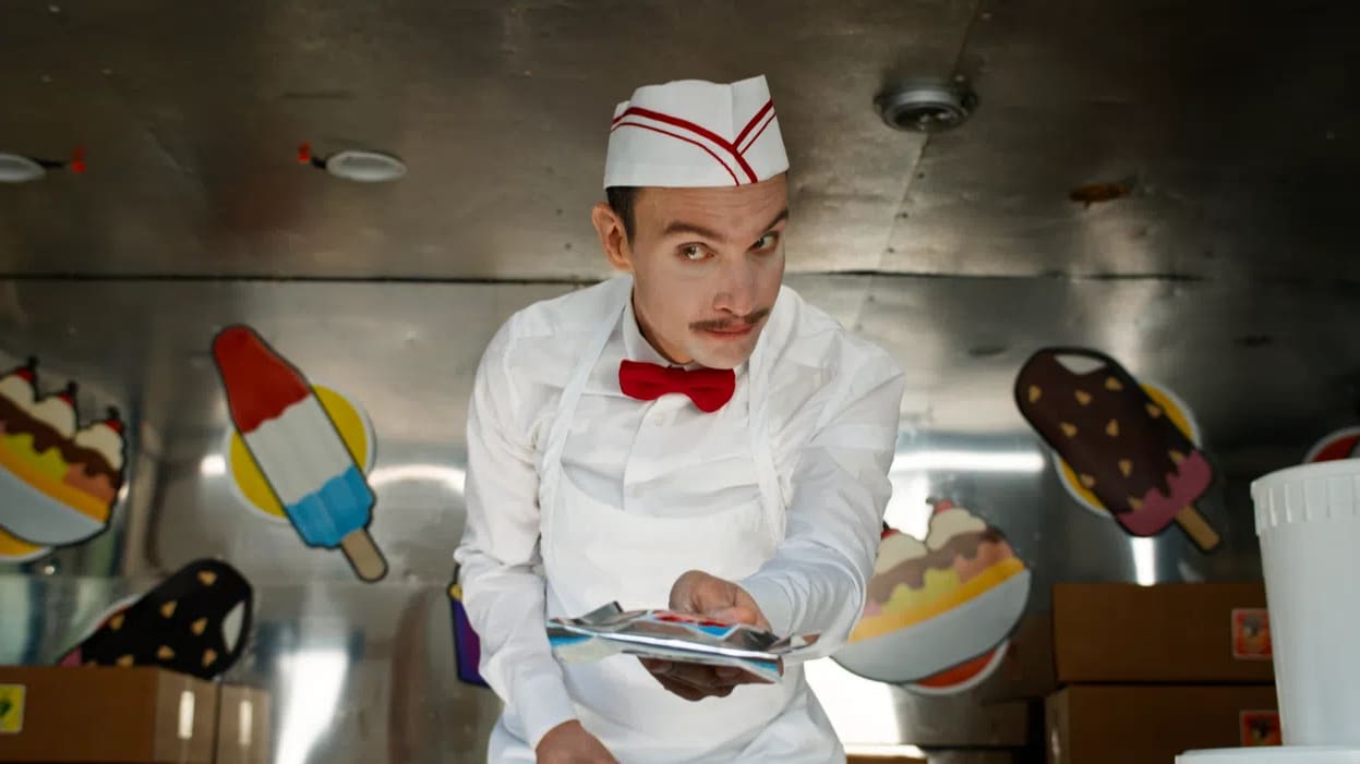 An official production promotional image from director Eli Roth's unrated horror-slasher film Ice Cream Man features actor Ari Millen portraying the menacing title character inside his dark, metallic ice cream truck. Leaning forward with an unsettling, wide-eyed stare and a sinister smirk, the slender vendor has a thin brown mustache and wears a vintage-style, crisp white uniform consisting of a long-sleeved button-up shirt, a matching white apron, a bright red bow tie, and a white paper soda jerk hat accented with red stripes. He extends his hand forward toward the camera, holding out a silver-wrapped frozen treat packet. In the softly blurred background, the interior of the food truck is visible, decorated with colorful, cartoonish graphics of frozen novelties including a rocket pop, an ice cream sundae bowl, and a chocolate-dipped ice cream bar with sprinkles, alongside stacked cardboard supply boxes sitting on the floor. 
