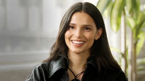 An elegant medium shot captures Inde Navarrette smiling warmly while looking directly forward against a softly blurred indoor backdrop. She has long, dark brown hair parted down the middle that drapes gracefully over her shoulders, framing her smooth complexion, defined cheekbones, and bright smile that reveals white teeth. Her ears are adorned with small gold hoop earrings, and she wears a collared black leather jacket over a matching top, styled with a simple dark cord necklace. The ambient lighting filters in from the side, illuminating her features and casting a gentle glow on the green leaves of a houseplant visible in the out-of-focus background.