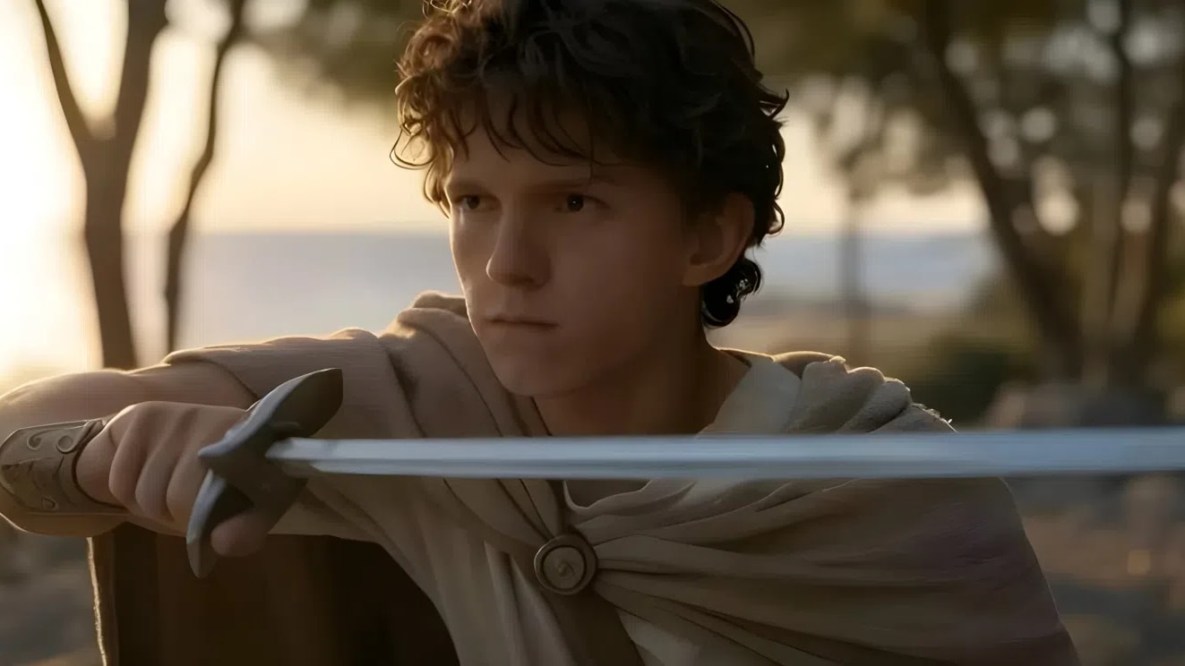 A wide, cinematic shot featuring actor Tom Holland as a young warrior in a historical or mythological setting. He is positioned in the lower-middle foreground, angled slightly to the right while looking intently toward the left. He has curly, dark brown hair and a serious, focused facial expression. He is holding a long, unsheathed silver sword horizontally in front of him with both hands, gripped near the dark hilt.He wears a light beige or cream-colored tunic paired with a draped brown cloak or cape secured at the center of his chest by a round, metallic medallion or brooch. On his right forearm, a dark leather bracer or cuff is visible. The background consists of a soft-focus, sun-drenched outdoor landscape with scattered, thin tree trunks and a hazy horizon line suggesting a coastal or Mediterranean environment during the golden hour. A small "SC" logo is visible in the bottom-left corner, and a faint Universal Pictures logo appears in the top-right corner.