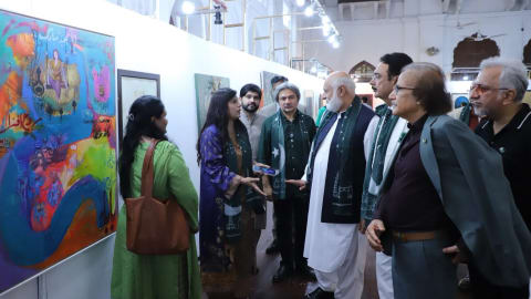 A vibrant indoor art exhibition features a diverse group of attendees gathered in a brightly lit, white-walled gallery space with high ceilings and traditional architectural balconies in the background. On the far left, a large, colorful, abstract painting filled with dense blue, orange, and green hues serves as a focal point. Several people, including a woman in a purple outfit and an older gentleman in a white shalwar kameez wearing a green scarf, stand in front of the artwork, actively engaged in a conversation while viewing the display. Other individuals, wearing formal attire or blazers, observe the gallery pieces under an overhead string of spotlights.