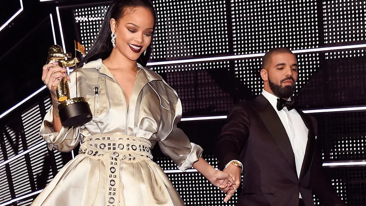 A medium-shot photograph showcases Rihanna and Drake on stage under bright concert lighting. On the left, Rihanna wears an elegant, collared, metallic silver-champagne gown structured like a trench coat, featuring a dramatically slit, flowing skirt and an intricately patterned, wide grommeted belt cinched at her waist. Her hair is styled sleek and straight behind her shoulders, and she wears dramatic dark maroon lipstick while looking downward with a smile. In her right hand, she holds a golden MTV "Moonman" trophy aloft, and with her left hand, she holds Drake’s hand. Drake stands to the right, dressed in a sharp black tuxedo with a crisp white shirt and a black bow tie, gazing toward her with a slight smile. The background features a large grid of geometric, black-and-white LED lighting, adding a modern, high-contrast texture to the scene.
