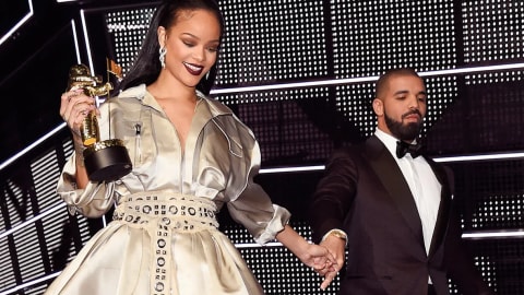 A medium-shot photograph showcases Rihanna and Drake on stage under bright concert lighting. On the left, Rihanna wears an elegant, collared, metallic silver-champagne gown structured like a trench coat, featuring a dramatically slit, flowing skirt and an intricately patterned, wide grommeted belt cinched at her waist. Her hair is styled sleek and straight behind her shoulders, and she wears dramatic dark maroon lipstick while looking downward with a smile. In her right hand, she holds a golden MTV "Moonman" trophy aloft, and with her left hand, she holds Drake’s hand. Drake stands to the right, dressed in a sharp black tuxedo with a crisp white shirt and a black bow tie, gazing toward her with a slight smile. The background features a large grid of geometric, black-and-white LED lighting, adding a modern, high-contrast texture to the scene.