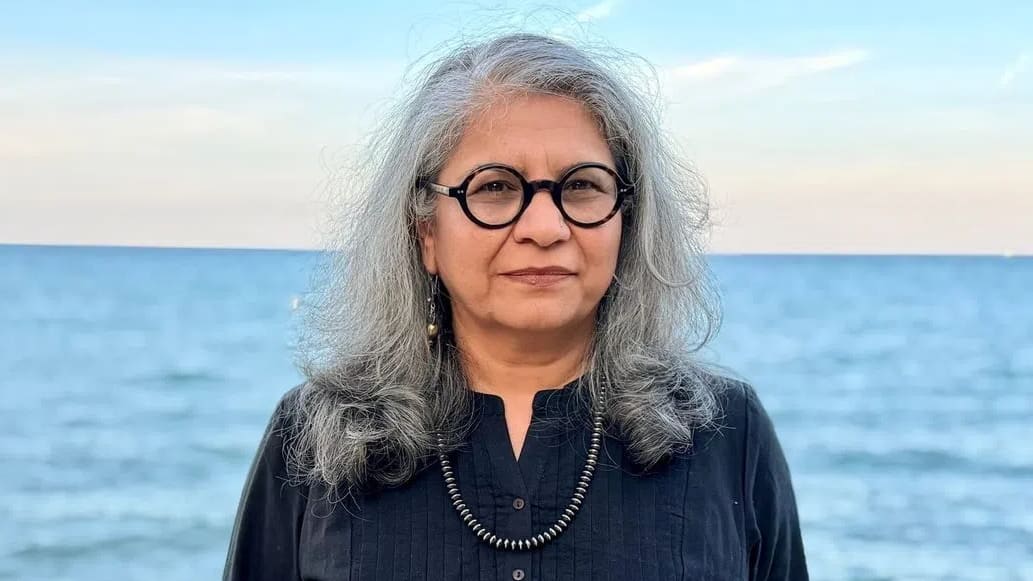 A medium close-up shot captures Dr. Savneet Talwar standing outdoors against a backdrop of a calm, blue sea meeting a light sky at the horizon line. She has shoulder-length, wavy grey hair parted to the side, and she wears round, thick-rimmed black glasses. Her expression is composed and neutral as she looks directly toward the camera. She is dressed in a dark black, long-sleeved button-up shirt featuring subtle vertical pleated detailing on the chest, complemented by a simple dark metallic beaded necklace. In the lower-left foreground, blurred rocky terrain lines the shore, establishing an outdoor coastal setting.