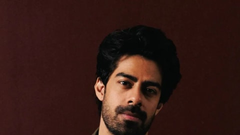 A medium shot of Zain Ahmad looking directly at the camera against a dark red paneled wall. He has thick black hair, a groomed beard, and a mustache. He is wearing an oversized olive green linen trench coat layered over a black button-up shirt that is slightly open at the collar, revealing a silver chain necklace. His hands are loosely placed near the pockets of his off-white or light beige linen trousers, creating a relaxed yet fashionable posture.