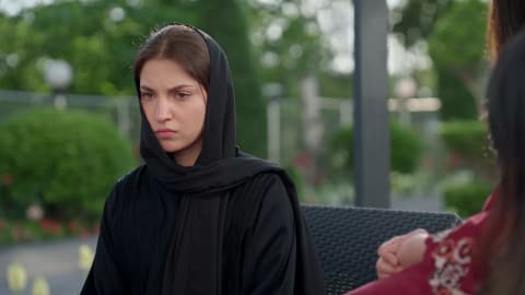 An intense close-up shot captures Pakistani actress Dananeer Mobeen portraying a moment of deep solemnity and emotional distress, her focused expression conveying heavy grief and determination. Clad in a traditional black abaya with a matching hijab pulled snugly over her head, she sits outdoors against a beautifully blurred background of lush green foliage and gardens. To the side, the soft-focus profile of another individual in a patterned maroon outfit hints at a tense, interpersonal conversation unfolding in an elegant open-air setting.
