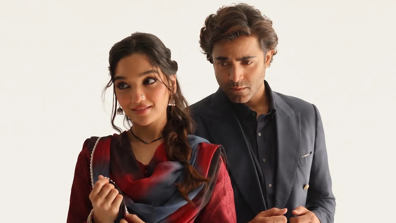 This promotional image features actors Sonya Hussyn and Sheheryar Munawar posing closely together for a contemporary Pakistani fashion collection campaign. A man and a woman pose closely together against a plain, light beige backdrop, showcasing elegant South Asian ethnic and formal attire. The woman on the left is captured from the waist up, looking slightly to her left with a gentle smile. Her dark hair is styled in a soft half-updo with loose, wavy tendrils framing her face, and she wears subtle traditional jewelry, including a thin black choker necklace, a nose stud, small metallic jhumka earrings, and a stack of silver bangles on her wrist. She is dressed in a deep crimson or maroon long-sleeved kurta paired with a lightweight chiffon dupatta draped over her shoulders. The dupatta features a striking tie-dye or ombre pattern with bold sweeps of dark gray, black, and maroon, finished with a delicate white lace or beaded border along the edges. To her right, the man stands slightly behind her, looking downward and to his left with a focused expression. He has wavy, short brown hair and wears a sophisticated charcoal gray or navy blue outfit consisting of a traditional straight-cut kurta shirt beneath a matching tailored, double-breasted blazer jacket with peak lapels and a white pocket square peeking out from the breast pocket. The lighting is soft and directional, casting gentle shadows that emphasize the rich textures of the fabrics and the warm tones of the scene.