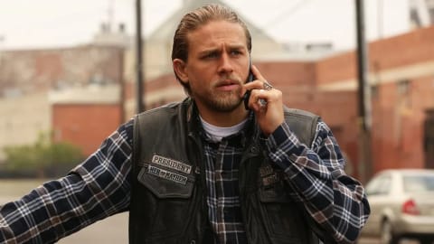 Jax Teller, the fierce president of the Sons of Anarchy Motorcycle Club, stands on a sun-drenched, gritty urban street while intensely focusing on a phone call. He sports his iconic biker look, donning a dark leather cuts vest layered over a blue, black, and white plaid flannel button-down shirt revealing a classic white crewneck undershirt beneath. His long blonde hair is slicked back neatly, complementing his rugged beard and mustache, while his left hand, adorned with prominent rings, holds the phone to his ear. The leather vest displays detailed, stitched club patches including "PRESIDENT" and "MEN OF MAYHEM" on the right breast, capturing his high-ranking status within the outlaw charter. In the soft-focus background, the hazy atmosphere of Charming features industrial buildings, sparse palm trees under an overcast sky, and a parked silver sedan, perfectly encapsulating the dramatic and tense mood of the television series.