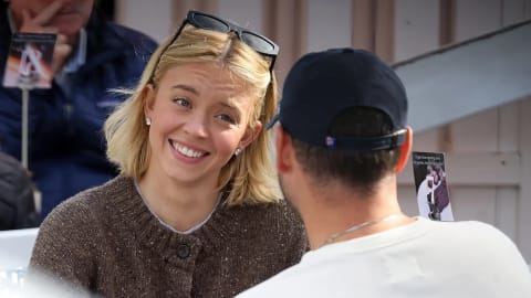 Sydney Sweeney and her partner, music executive Scooter Braun, are pictured enjoying an intimate moment together outdoors. A candid, medium close-up shot capturing actress Sydney Sweeney sharing a warm, smiling interaction with Scooter Braun. Sweeney, situated on the left half of the frame and facing slightly right, smiles brightly with her teeth showing, looking toward Braun with a cheerful and affectionate expression. She has blonde, shoulder-length bobbed hair parted down the middle, with a pair of black sunglasses resting on top of her head. She wears a chunky, dark brown knit button-up sweater cardigan over a simple white top, complemented by small, elegant stud earrings. Braun is positioned on the right half of the frame, captured from a back-facing profile perspective that leaves his face obscured from view. He wears a clean white long-sleeved crewneck t-shirt and a dark navy blue baseball cap worn forward. The background features light-colored rustic wooden paneling, a hint of an outdoor cafe chair back with a blue and white woven pattern, and blurry figures in the distant left, emphasizing the focus on the couple's intimate dynamic.