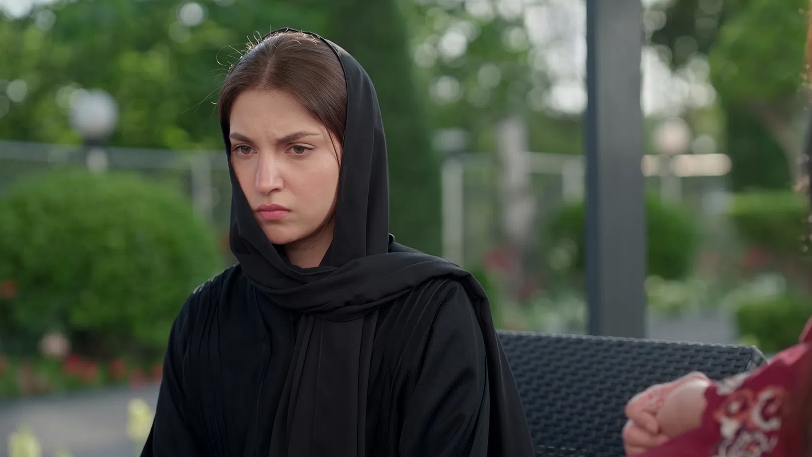 An intense close-up shot captures Pakistani actress Dananeer Mobeen portraying a moment of deep solemnity and emotional distress, her focused expression conveying heavy grief and determination. Clad in a traditional black abaya with a matching hijab pulled snugly over her head, she sits outdoors against a beautifully blurred background of lush green foliage and gardens. To the side, the soft-focus profile of another individual in a patterned maroon outfit hints at a tense, interpersonal conversation unfolding in an elegant open-air setting.