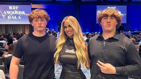 Kim Zolciak stands proudly between her two teenage sons, KJ and Kash Biermann, while attending the B2C Awards Gala Ceremony. The reality television personality wears a fitted, shimmering metallic dark grey gown featuring a mock neck and an asymmetrical thigh-high slit that highlights her figure. Her long blonde hair is styled in loose, voluminous waves draping over her shoulders. Flanking her on the left, one son wears a simple black crewneck t-shirt paired with matching black trousers, while the other son stands to her right in a black long-sleeved button-down shirt, a silver cross necklace, and matching black dress pants as he flashes a peace sign toward the camera. They pose together in a large, carpeted event hall filled with rows of seated attendees beneath bright blue overhead event lighting.