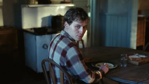 A medium shot captures actor Fred Hechinger as Mike sitting at a rustic wooden dinner table inside a dimly lit cabin kitchen, glancing over his shoulder with an intense, wary expression. He wears a brown, beige, and white plaid flannel shirt and holds a fork in his hand over a ceramic plate, while a half-empty glass of water sits next to a wooden cutting board with sliced bread nearby. The background is softly blurred, revealing a vintage white kitchen cabinet unit and a dark stove cast in shadows, emphasizing the somber and rustic 1950s period setting.