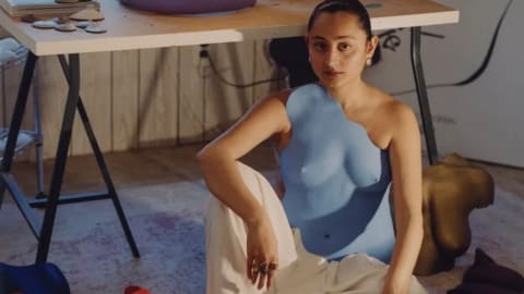 In this portrait, the Pakistani artist and fashion designer Misha Japanwala sits on the floor of her art studio surrounded by several of her signature, handcrafted body-cast sculptures. She is positioned centrally, looking directly at the camera while wearing white, wide-leg trousers and a striking, asymmetrical sky-blue resin breastplate molded to her torso. Behind her stands a light-wood work desk covered with art supplies, including a jar of paintbrushes and candles, and topped by a large, lavender-colored sculptural torso. Propped against the wall behind the desk is a large canvas featuring a minimalist black-and-white calligraphic silhouette drawing. Scattered around her on the floor are additional sculpted anatomical forms in bold shades of red, dark blue, gold, and dark purple, highlighting her unique creative practice that explores themes of bodily autonomy and the deconstruction of shame.