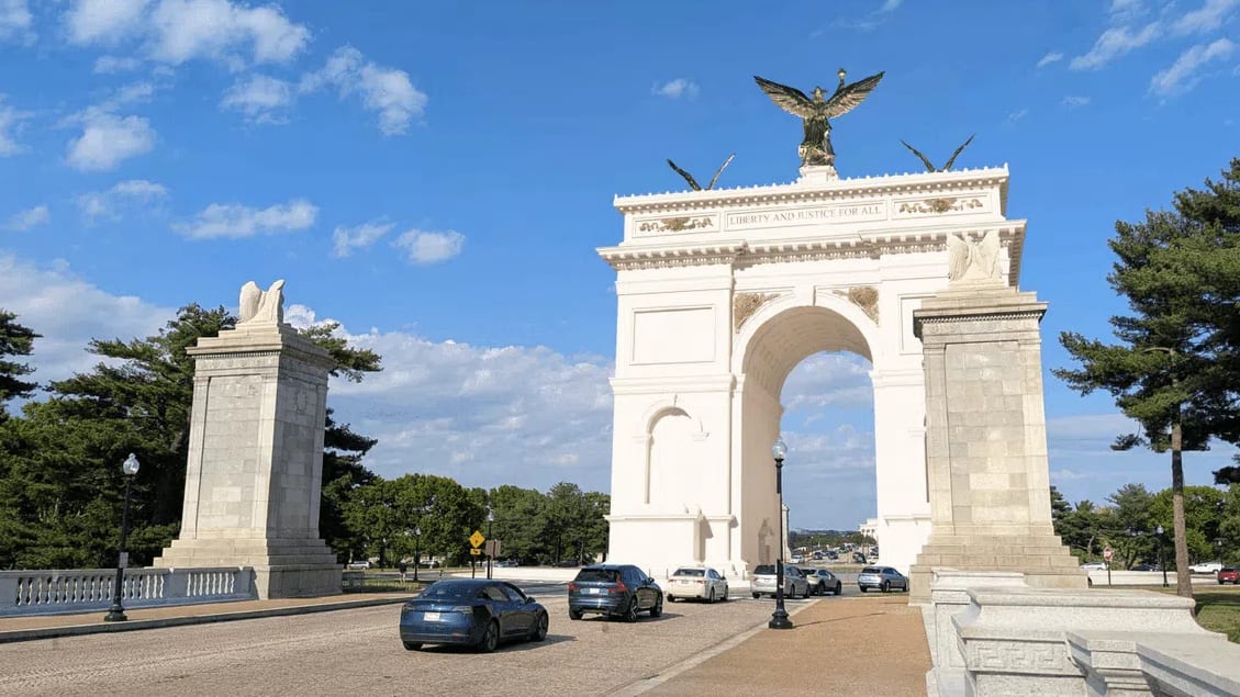 This digital rendering depicts the proposed neoclassical United States Triumphal Arch, an immense white stone monument spanning a multi-lane roadway filled with passing sedans. Positioned along the Arlington Memorial Bridge approach near Memorial Circle, the structure features a large central barrel-vaulted arch flanked by detailed architectural columns and decorative side niches. The top frieze of the main structure is inscribed with the phrase "LIBERTY AND JUSTICE FOR ALL" and is crowned by three prominent golden sculptures, including a large, central winged figure holding a torch and crown aloft, flanked by two smaller winged figures. Separate, heavy stone pylons with sculpted eagle motifs stand on either side of the entrance lane, framing the gateway against a bright blue sky filled with scattered white clouds. 