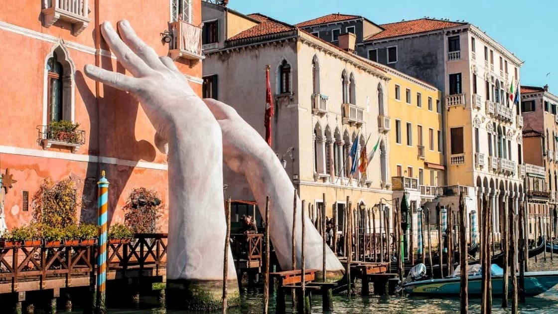The monumental sculpture shown is "Support" (2017) by contemporary Italian artist Lorenzo Quinn. This detailed photograph captures a large-scale public art installation in Venice, featuring two massive, white, child-like hands emerging from the deep green waters of the Grand Canal. The hands are positioned as if they are literally supporting and bracing the historic, salmon-colored facade of the Ca' Sagredo Hotel. One hand grips the corner of the ornate building while the other reaches up against its side, set against a backdrop of sunlit Venetian architecture and traditional wooden mooring poles lining the canal. 