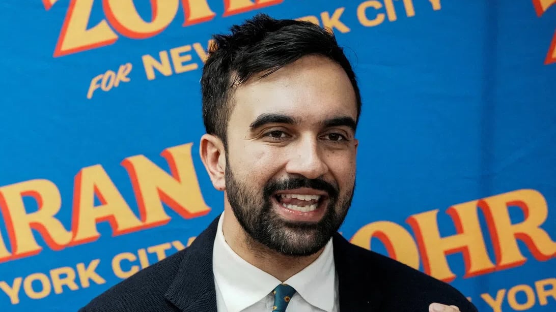 A man with short dark hair and a beard stands in front of a blue background that features a repetitive orange and yellow logo reading "ZOHRAN FOR NEW YORK CITY." He is wearing a dark blazer over a white collared shirt paired with a green tie that has a small, repeating patterned design. He is captured mid-speech, looking to his right with his mouth open and his left hand raised, fingers partially curled, as if gesturing to emphasize a point.