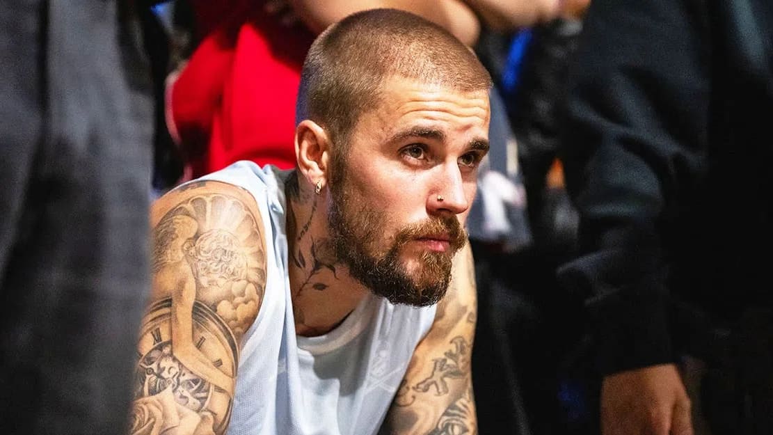 A close-up shot captures Justin Bieber from the chest up, leaning slightly forward with a pensive and focused expression while looking toward the right. He features a buzz-cut hairstyle, a well-groomed full beard, a mustache, a small hoop earring in his right ear, and a subtle nose stud. He is wearing a light blue sleeveless jersey, which fully exposes his arms and neck to reveal extensive, intricate black-and-grey ink tattoos, including an angel and a clock face design on his right shoulder and bicep. The foreground and background are softly blurred, showing glimpses of dark clothing from individuals standing nearby, creating a shallow depth of field that keeps the central focus entirely on his contemplative demeanor.