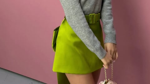 A young woman with vibrant pink hair and bangs poses against a pink background, wearing a grey long-sleeved sweater with a large white flower pin on the shoulder and a structured lime-green belted miniskirt while holding a small gold chain handbag. The image showcases a vibrant and stylish fashion portrait set against a solid, matte pink background. A young woman with long, straight pink hair and a clean fringe leans gracefully to her left, looking directly into the camera. She wears a long-sleeved, grey knit sweater adorned with a large, white fabric flower pinned to her left shoulder. The sweater is tucked into a bright, structured lime-green miniskirt featuring a matching fabric belt with a square buckle. The skirt includes a structured panel or bow detailing extending down the side and back. Her left hand rests near the belt, while her right hand gently holds a small, embellished gold chain purse.
