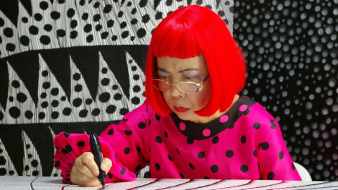 The image shows the famous contemporary Japanese artist Yayoi Kusama working diligently at a desk, deeply focused on drawing intricate patterns with a black marker on a large canvas. She is instantly recognizable by her vibrant, bright red bob haircut with straight-across bangs, gold-rimmed reading glasses, and dark eyeshadow. She wears a striking hot pink blouse covered in large black polka dots, featuring a black collar and long sleeves with gathered, elasticated cuffs. Her hand holds the marker firmly as she guides it across a surface densely covered in her signature fine lines and repetitive dot motifs. The background behind her is a visually complex, high-contrast environment filled with her artwork, displaying large black and white geometric shapes, alternating fields of dense polka dots, and vertical patterns that create an immersive, hallucinatory atmosphere characteristic of her iconic style.A high-contrast photograph of artist Yayoi Kusama in her signature bright red bob and polka-dot blouse creating detailed artwork.