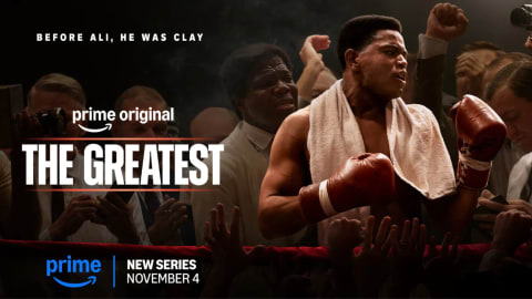 An official promotional poster for the Amazon Prime Video biographical drama series The Greatest, an authorized scripted chronicle showcasing the life and legacy of boxing champion, civil rights icon, and global humanitarian Muhammad Ali. Set against a dramatic, dimly lit boxing ring background shrouded in subtle smoke, the scene centers on actor Jaalen Best capturing a triumphant, high-intensity moment in his portrayal of a young Cassius Clay. He stands chest-baring with a white terry cloth towel slung loosely over his neck, his muscles defined and glistening under a focused overhead spotlight as his face twists into a fierce, mid-battle or celebratory shout directed away from the camera. His hands are raised in red boxing gloves, flanked on either side by a crowded arena of black-and-white toned spectators, press photographers lifting vintage cameras, and supporting figures closing in around the ropes. On the left side, bold, clean white text layers over the crowd reading "BEFORE ALI, HE WAS CLAY" stacked above a glowing, stylized blue "prime original" logo framework that underlines the main title, "THE GREATEST", written in a massive, clean white sans-serif block font. Along the bottom edge, a vibrant blue Amazon Prime logo pairs with a bold sans-serif title card announcing the project as a "NEW SERIES NOVEMBER 4", anchoring the cinematic, vintage-toned promotional imagery.