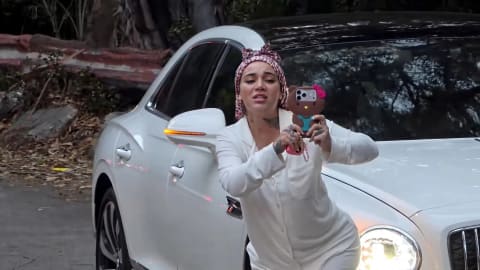 American media personality and rapper Danielle Bregoli, widely recognized as Bhad Bhabie, leans dynamically forward against a luxurious white Bentley Flying Spur while aggressively aiming her smartphone outward during a highly publicized confrontation in Hollywood. She is dressed in an off-white, long-sleeved loungewear set paired with a pink-and-white leopard-print headscarf wrapped tightly over her hair. Her expression is visually tense, marked by furrowed brows and parted lips as she fiercely records or gestures toward an off-screen group of live streamers. Her smartphone is prominently enclosed in a bulky, distinctive brown-and-pink Hello Kitty protective phone case. The luxury car behind her sits angled on a paved asphalt roadway, showing illuminated circular headlights and a glowing orange side-marker signal mirroring the chaotic, high-visibility energy of the scene. In the dimly lit background, natural foliage, overhanging branches, and a concrete roadside barrier provide a stark, unrefined contrast to the sleek design of the premium sedan.