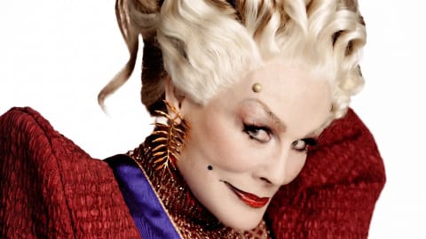 Glenn Close commands the frame with a wicked, theatrical intensity as Drusilla Sickle, showcasing a striking villainous look complete with an elaborate platinum blonde wig styled in structured vintage waves, dramatic arched eyebrows accented by a gold micro-stud, and a sharp, glossy red lip featuring a defined dark outline. Her piercing gaze is directed sharply to the side while wearing an oversized, structural red textured collar that frames her face, paired with a matching gold fern-like statement earring that perfectly complements her sinister, aristocratic aesthetic. Glenn Close transformed as the character Drusilla Sickle in The Hunger Games: Sunrise on the Reaping.
