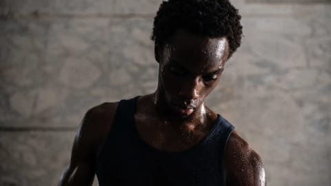 Kai Cenat commands attention in a striking promotional image for the VIVET New York Fashion Week (NYFW) showcase, capturing a moment of raw, cinematic intensity. The internet personality and fashion icon is captured from the chest up against a textured, neutral-toned concrete backdrop that enhances the gritty, high-contrast atmosphere of the photograph. Looking downwards with a pensive, focused expression, his face and dark, coiled hair are dramatically highlighted by a sharp key light that catches the sheen of heavy sweat beads covering his skin. He wears a form-fitting, dark ribbed tank top that emphasizes his athletic frame and glistening shoulders, blending sportswear aesthetics with high-fashion drama. The low-key, moody lighting casts deep shadows across his features, shifting the focus to the intense texture of the sweat and the subtle contours of his expression to create a powerful, atmospheric portrait of athletic endurance and modern style.