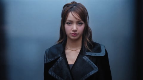 The selected region features K-pop global superstar LISA looking directly into the camera during a promo clip for her major brand crossover campaign. Her hair is styled in a sleek half-up look with casual, face-framing brown bangs draped gently over her forehead, complementing her clean wings of dark eyeliner and a subtle lip color. She is wearing a structured black statement jacket that stands out against a cool, foggy blue background, featuring prominent wide lapels with distinct trim textures and a thin gold chain resting around her collarbone. Following her initial global ambassador campaign for the PS5, this sleek aesthetic aligns with the official launch of the limited-edition custom DualSense controller and exclusive apparel merch line designed directly by the artist.