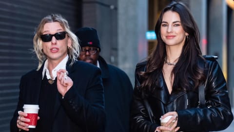 An eye-level, medium shot captures three individuals walking down a city street flanked by industrial buildings.On the left, a person with blonde, shoulder-length hair wears black sunglasses, a long black overcoat, a white collared shirt under a black sweater, and dark, pinstriped trousers. They hold a white paper coffee cup with a red sleeve in their right hand, capturing a candid, mid-speech expression.On the right, a woman with long, dark brown hair smiles forward while walking. She is dressed in a glossy black leather trench coat over dark gray trousers, holding a white coffee cup in her left hand.Slightly behind them and positioned in the center, a third individual wears a black beanie, glasses, and a dark coat, looking toward the left.The background features a gray, corrugated metal garage door on the left and a stone building facade on the right, casting soft, diffused daylight over the scene.
