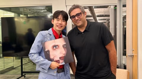 A brightly lit modern office setting showcases two men posing side-by-side. On the left, a smiling young East Asian man with short dark hair wears an elaborate light blue Western-style suit adorned with white floral embroidery along the lapels and sleeves, styled over a vibrant red button-down shirt. He proudly holds a copy of Lady Gaga's vinyl album, Mayhem, featuring a prominent close-up artwork of fragmented faces. To his right, Michael Polansky stands casually with one hand in his pocket. He is a middle-aged man with salt-and-pepper hair, wearing rectangular black-rimmed glasses and a simple short-sleeved black polo shirt. Large glass window panels and corporate office elements fill the background.