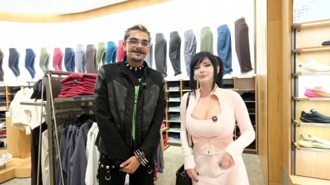 An overhead shot captures social media personalities Pinkchyu and N3on standing side-by-side inside a brightly lit Lululemon retail store, surrounded by displays of athletic apparel and footwear. On the left, N3on stands with his hands clasped forward, sporting a black jacket over a green graphic shirt, edgy spiked jewelry, and a thin, stylized mustache. To his right, Pinkchyu smiles toward the camera, dressed in a form-fitting, pale pink athletic zip-up jacket and matching top, with her dark hair styled in high pigtails. The background features neat rows of colorful leggings displayed on mannequins, stacks of folded hoodies, and a wooden shelf on the right showcasing various running shoes.