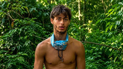 A medium close-up shot captures Stavros Floros, a contestant from the 13th season of the reality TV show Survivor Greece, standing shirt-less in front of a dense, lush green jungle backdrop. He is looking slightly off-camera to his right with an open-mouthed, intense expression. He wears a light blue Survivor buff rolled around his neck like a collar, with a dark cord necklace holding a small cross pendant dangling down his chest. The bright, natural sunlight filters through the green foliage, illuminating his disheveled brown hair, stubble, and sun-tanned shoulders while casting soft shadows across his torso.