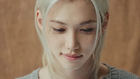 The image shows a close-up portrait of a young individual with striking features, captured from a slightly elevated angle as they look downward with a soft, serene expression. Their long, straight, platinum-blonde hair is parted down the middle, framing a face characterized by pale skin, dark, well-defined eyebrows, and prominent, natural freckles dusting the cheeks and bridge of the nose. Their eyes are cast down, and a gentle, subtle smile plays on their pink lips, giving the portrait a contemplative and calm demeanor. The lighting is soft and even, highlighting the smooth texture of the skin and the individual strands of hair, while the background is a muted, warm beige tone that keeps the focus entirely on the subject. In the lower-left corner, the vibrant blue "JYP" logo and white "OFFICIAL" text are visible, with the words "Slow It Down" written in a clean white serif font just above them.