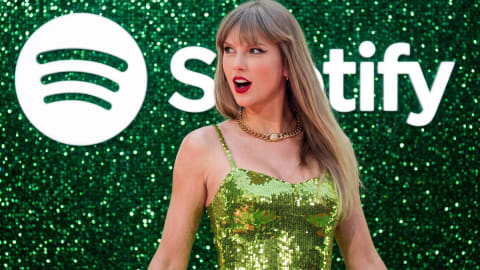 Taylor Swift sports a vibrant, lime-green sequined dress paired with a chunky gold chain necklace as she poses dynamically in front of a sparkling green backdrop adorned with the Spotify logo. Her long blonde hair is styled straight with signature full bangs, and her striking makeup features a sharp, black winged eyeliner and a bold, classic red lip. Her expression is playful and animated, captured mid-glance to the side with an open-mouthed, surprised look that adds a lively, candid energy to the red carpet event. The dress's thin spaghetti straps and scoop neckline showcase the prominent necklace, while the textured, shimmering wall behind her reflects countless pinpricks of light, matching the glitter of her outfit and amplifying the festive, high-profile atmosphere of the occasion.