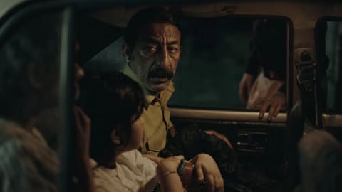 A dimly lit, cinematic film still captured from inside a vintage automobile at night showcases a tense and suspenseful sequence. The focal point of the composition is a middle-aged South Asian man with a prominent mustache and an anxious expression, dressed in a light-colored button-up shirt, looking warily out the open driver-side window. A hand rests heavily on the car's window frame from the outside, belonging to an unseen figure standing in the darkness, hinting at an active interrogation or confrontation. In the foreground, the back of a young child's head with dark hair is visible, seated next to another partially obscured passenger on the left, capturing a quiet, stressful moment shared within the vehicle's cramped interior.